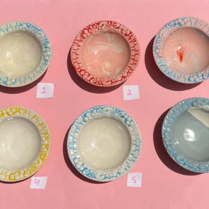May include: Six small, round ceramic bowls with floral patterned rims. The bowls are in various colors, including yellow, blue, red, and pink, with a light-colored interior. Each bowl is numbered on a small white tag.