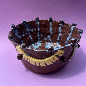 May include: A handmade ceramic bowl with a dark brown exterior and a blue and brown interior. The bowl has a unique shape with a wavy rim and is decorated with yellow and brown accents. Small brown spheres adorn the rim.