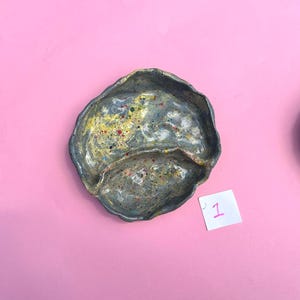 May include: A handmade ceramic dish with a unique, organic shape. The dish is divided into two sections and features a speckled, mottled glaze in shades of gray, yellow, and red. The dish sits on a pink background. A small white square with the number "1" is in the image.