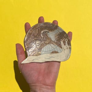 May include: A ceramic snail-shaped dish with a brown and cream-colored shell and a cream-colored body. The dish has two small antennae and is set against a bright yellow background.