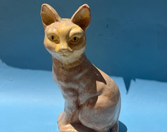 May include: A ceramic cat figurine in a seated position. The cat has tan fur with darker brown markings and yellow eyes. The ears are pointed, and the cat is sitting upright. The figurine is on a blue surface.