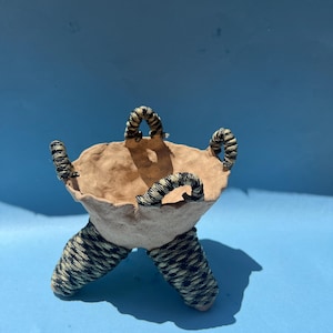 May include: A handmade, beige-colored bowl with four handles and two legs, all wrapped in a black and white patterned material. The bowl is set against a solid blue background, showcasing its unique design and texture.