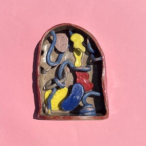 Puede incluir: Una pieza de arte de cerámica con borde rojo e interior beige. El interior presenta formas abstractas en azul, amarillo, rojo y rosa. La pieza está sobre un fondo rosa.