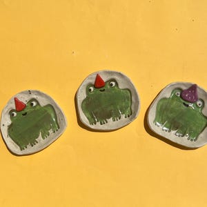 Può includere: Tre piatti in ceramica a forma di rana, con base grigio chiaro e dettagli di rana verde. Ogni rana indossa un cappello da festa, uno rosso, uno viola e uno rosso. I piatti sono su sfondo giallo.