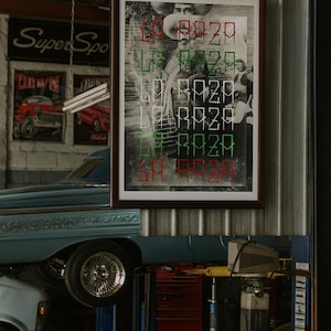 May include: A framed black and white print with the text "LA RAZA" in red, green, and white. The artwork hangs in a garage setting, with a classic car on a lift in the foreground and vintage car posters in the background.