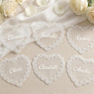 Può includere: Sei segnaposto a forma di cuore con bordi in pizzo bianco e nomi in elegante scrittura: Eleanor, Isabella, Sophia, Olivia, Charlotte e Amelia. Le carte sono disposte su una superficie color crema con rose bianche.