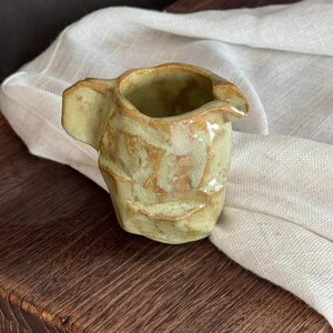Puede incluir: Una pequeña jarra de cerámica hecha a mano con una textura irregular y arrugada, cubierta por un esmalte brillante en tonos verde claro y bronceado. El recipiente cuenta con un asa corta y un pequeño pico vertedor integrado en el borde.