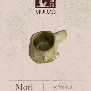 May include: A small, handcrafted coffee cup with an irregular shape and a pale green glaze. The cup has a small handle and is labelled "Mori coffee cup" below the Mudzō logo, which features a bird and Japanese characters.