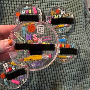 May include: A collection of clear, round resin coasters with school-themed embellishments. Each coaster features miniature pencils, paperclips, and glitter, with the titles "MRS", "MS", and "MR" in colorful letters. The coasters are designed for teachers.