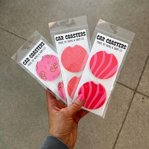 May include: Three car coasters in clear packaging. Each coaster has a different pink design: cherries, leopard print, and wavy stripes. The packaging reads "CAR COASTERS MADE BY BRAG & BRIM CO."