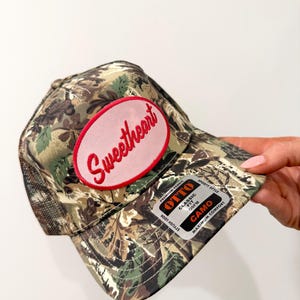 Può includere: Cappellino da baseball mimetico con una toppa ovale rosa con la scritta "Sweetheart" in corsivo rosso. Il cappellino ha un retro in rete e una visiera curva. L'etichetta recita "OTTO CAMO".