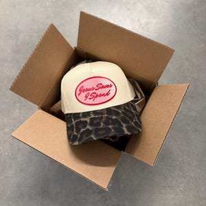 May include: A beige baseball cap with a leopard print brim and the text "Jesus Saves I Spend" in a red oval. The cap is inside an open cardboard box, suggesting a recent purchase or delivery.