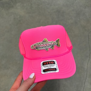 May include: A neon pink trucker hat featuring a foam front panel and a mesh back. An embroidered patch of a rainbow trout is centered on the front. A white rectangular sticker on the brim reads OTTO WORLD FAMOUS FOAM FRONT OSFM TRUCKER.
