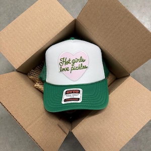 Könnte beinhalten: Eine grün-weiße Trucker-Cap mit einer Schaumstoff-Frontpartie und einem hellrosa herzförmigen Aufnäher. Der Text "Hot girls love pickles" ist in grüner Schreibschrift auf das Herz gestickt. Ein Otto-Markenaufkleber befindet sich auf dem Schirm.