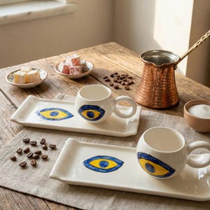 Puede incluir: Dos tazas de café de cerámica blanca con un diseño de ojo azul y amarillo, cada una sobre una bandeja rectangular a juego. También se ve una cafetera de cobre, pequeños platos de delicias turcas y granos de café.