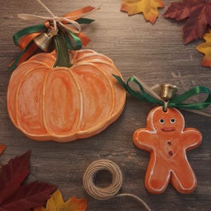 Könnte beinhalten: Orangefarbene Keramik-Kürbis- und Lebkuchenmann-Ornamente mit weißen Akzenten und kleinen Glocken. Der Kürbis hat einen grünen Stiel und orangefarbene und grüne Bänder. Der Lebkuchenmann hat ein lächelndes Gesicht und rote Knöpfe. Eine Garnrolle ist ebenfalls vorhanden.
