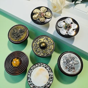 May include: A selection of decorative cabinet knobs with diverse designs. The knobs showcase floral, butterfly, and abstract patterns in gold, silver, and brown hues. Some knobs have a wooden base, others a metal base.