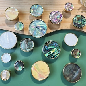 May include: A collection of decorative cabinet knobs with iridescent abalone shell centers and gold-toned rims. The knobs vary in size and color, with shades of blue, green, purple, and white. They are arranged on a green and wood-toned surface.