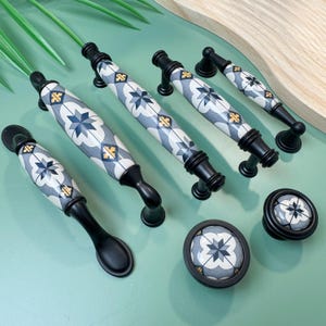 May include: A collection of cabinet hardware, including handles and knobs. The handles have a black base and a ceramic center with a blue, white, and yellow geometric pattern. The knobs have a matching design. The hardware is displayed on a light green surface.