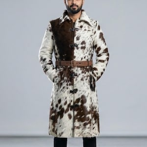 May include: A long coat with a cowhide pattern in brown and white. The coat has a brown leather belt and a button closure. The collar is a contrasting brown color. The coat has pockets and is knee-length.