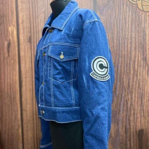 May include: A blue denim jacket with white stitching and gold buttons. The jacket has two chest pockets and a Capsule Corp patch on the sleeve. The jacket is displayed on a mannequin.
