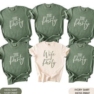 Könnte beinhalten: Sechs T-Shirts, fünf in Moosgrün und eines in Elfenbein. Die moosgrünen Shirts haben "THE Party" in weißer Schrift aufgedruckt. Das elfenbeinfarbene Shirt trägt "Wife of the Party".