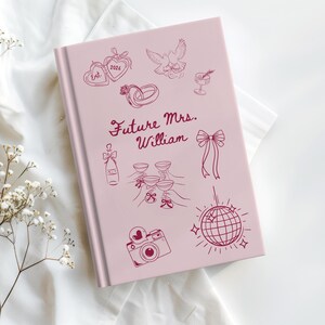 Op de afbeelding: Een roze notitieboek met de tekst "Future Mrs. William" en illustraties van huwelijkssymbolen zoals ringen, champagneglazen en een discobal. Het hardcover notitieboek is bedoeld voor bruiloftplanning.