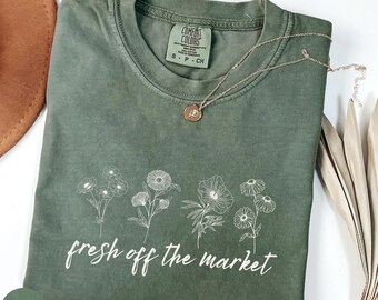 Op de afbeelding: Een mosgroen t-shirt met een bloemenontwerp en de tekst "fresh off the market". Het shirt heeft een ronde hals en korte mouwen. Een gouden ketting met een hanger ligt op het shirt. Het shirt is op een wit oppervlak gevouwen.