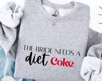 Op de afbeelding: Een lichtgrijs sweatshirt met de tekst "THE BRIDE NEEDS A diet Coke" in zwart en rood. Een zilveren ketting met een zonvormige hanger ligt op het shirt. Het sweatshirt is op een blauwe spijkerbroek gevouwen.