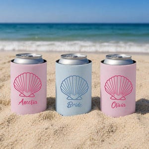 Op de afbeelding: Drie blikjeskoelers op een zandstrand. Twee roze koelers met een schelpmotief en de namen "Amelia" en "Olivia". De derde koeler is lichtblauw met een schelpmotief en het woord "Bride".