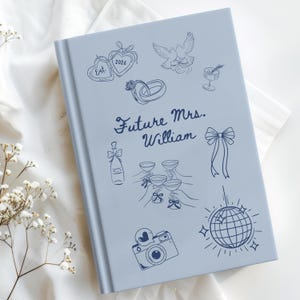 Op de afbeelding: Een lichtblauw trouwgastboek met verschillende illustraties in lijntekening, waaronder harten, duiven, champagneglazen en een discobal. De tekst "Future Mrs. William" is geschreven in elegant schrift.
