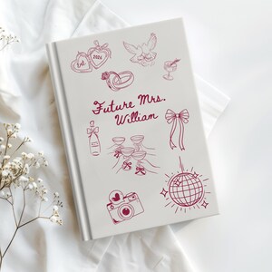 Op de afbeelding: Een wit notitieboek met een bordeauxrode illustratie van bruiloft-thema-iconen, waaronder ringen, champagneglazen en een discobal. De tekst "Future Mrs. William" is geschreven in bordeauxrode letters.