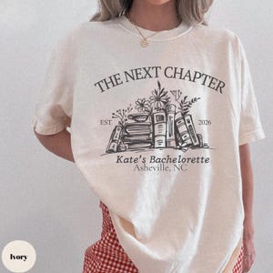 May include: Ivory t-shirt featuring the text "THE NEXT CHAPTER" above a graphic of books and flowers, with "Kate's Bachelorette Asheville, NC" below. The shirt also has the text "EST. 2026".
