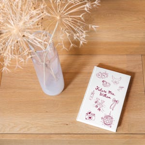 Op de afbeelding: Een wit notitieboek met de tekst "Future Mrs. William" en diverse illustraties met een bruiloftthema in bordeauxrode inkt. Het notitieboek ligt op een houten oppervlak naast een transparante vaas met gedroogde bloemen.