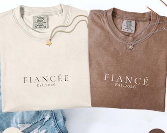 Op de afbeelding: Twee T-shirts, één ivoor en één espresso, met de woorden "FIANCÉE" en "FIANCÉ" erop gedrukt. Elk shirt heeft "Est.2026" eronder gedrukt. Gouden en zilveren kettingen zijn over de shirts gedrapeerd. Het ivoorkleurige shirt is maat XL-TG en het espresso-shirt is maat S-CH.