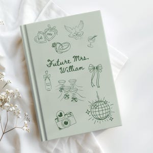 Op de afbeelding: Een lichtgroen gastenboek voor een bruiloft met handgetekende illustraties van harten, duiven, champagneglazen en een discobal. De tekst "Future Mrs. William" is geschreven in een elegant lettertype. Het boek is gesloten en ligt op een wit oppervlak.