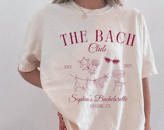 May include: Ivory-colored t-shirt with a crimson print. The design includes the text "THE BACH Club" and "Sophia's Bachelorette MALIBU, CA". Graphics of seashells, sunglasses, and swimwear are also featured. The shirt is a relaxed fit.