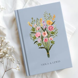 May include: A pale blue journal featuring a watercolour floral bouquet. The bouquet includes pink, yellow, and orange flowers with green leaves, tied with a gold ribbon. The text "VIOLA & LEWIS" is printed at the bottom.