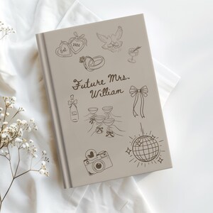 Op de afbeelding: Een lichtgrijs notitieboek met de tekst "Future Mrs. William" en diverse huwelijksthema-illustraties. De cover toont een hart, ringen, een duif, champagne, glazen, een strik, een camera en een discobal.
