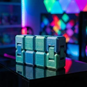 Puede incluir: Un juguete fidget impreso en 3D en tonos azules y verdes se encuentra sobre una superficie negra. El juguete está compuesto por bloques rectangulares entrelazados. El fondo presenta luces LED coloridas y una imagen borrosa de una pantalla de ordenador.