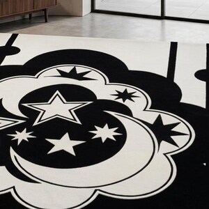 May include: A monochrome rug with a celestial design. The rug has a cream background with a large black cloud-like shape in the centre, filled with stars and crescent moons. The rug is rectangular in shape.