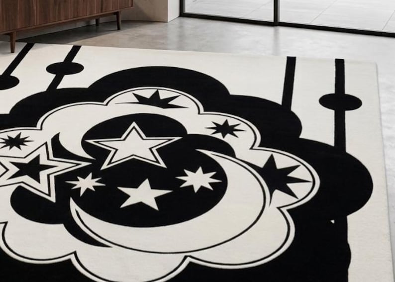 May include: A monochrome rug with a celestial design. The rug has a cream background with a large black cloud-like shape in the centre, filled with stars and crescent moons. The rug is rectangular in shape.