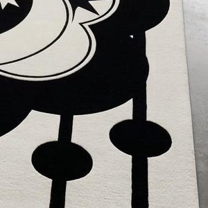 May include: A black and white rug with a celestial design. The rug features a large, stylized moon and star pattern in black against a white background. The design includes a series of black, circular shapes connected by vertical lines.