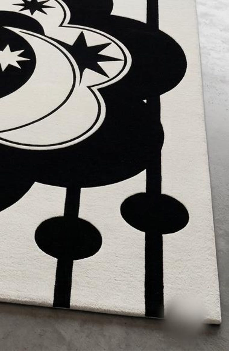 May include: A black and white rug with a celestial design. The rug features a large, stylized moon and star pattern in black against a white background. The design includes a series of black, circular shapes connected by vertical lines.