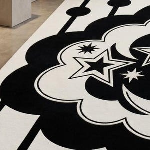 May include: A close-up of a black and white patterned rug. The rug features a repeating design of stylized clouds, stars, and crescent moons. The design is in black against a white background. The rug is made of a soft, plush material.