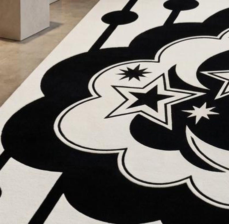 May include: A close-up of a black and white patterned rug. The rug features a repeating design of stylized clouds, stars, and crescent moons. The design is in black against a white background. The rug is made of a soft, plush material.