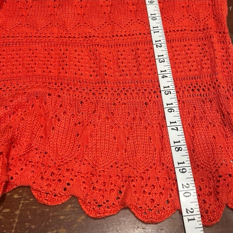 May include: Close-up of a bright orange knitted garment. The fabric has a detailed openwork design with a scalloped hem. A measuring tape shows the garment is approximately 21 inches long.