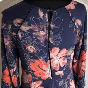 K&ouml;nnte beinhalten: Marineblaues Langarmkleid mit floralem Muster in Korallen-, Rosa- und Wei&szlig;t&ouml;nen. Das Kleid hat einen Schl&uuml;sselloch-Ausschnitt und dekorative N&auml;hte an den Schultern. Das florale Design ist eine Mischung aus gro&szlig;en und kleinen Bl&uuml;ten.