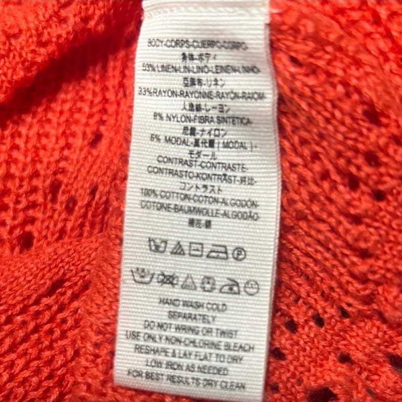May include: Close-up of a tag with fabric composition and care instructions. The tag is white with black text and is attached to a bright orange knit fabric. The tag lists the fabric content as linen, rayon, nylon, modal, and cotton.