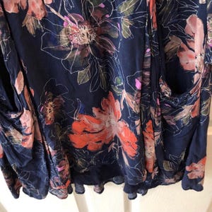 K&ouml;nnte beinhalten: Ein marineblaues Kleidungsst&uuml;ck mit floralem Muster. Das Design zeigt gro&szlig;e, stilisierte Bl&uuml;ten in Korallen-, Rosa- und Lilat&ouml;nen mit wei&szlig;en Konturen. Der Stoff wirkt weich und flie&szlig;end, was auf ein Kleid oder ein Oberteil hindeutet.